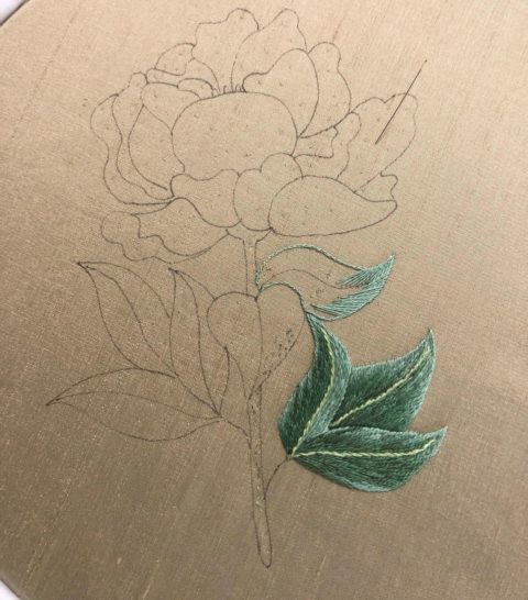 a hand-drawn outline of a peony on beige silk with one leaf embroidered in green