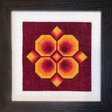 A 1970s inspired cross stitch of a four sided geometric flower stitched in reds and yellows on a maroon background and displayed in a black wood frame