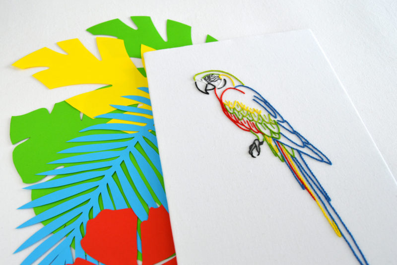 Embroidery of a colorful macaw resting on top paper cutouts of tropical leaves