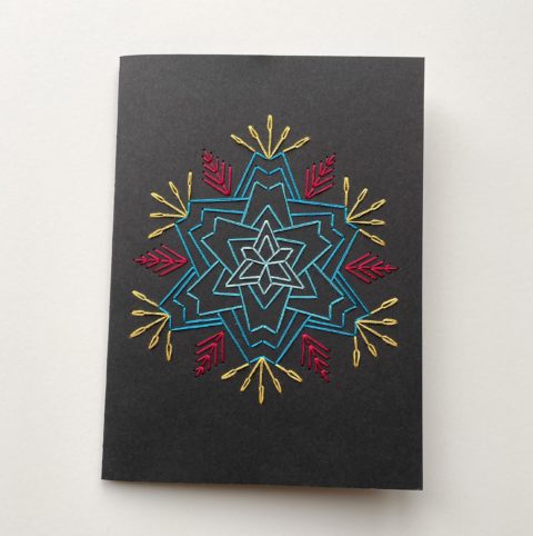 A star-shaped flower in blue, raspberry, and yellow, stitched on a black greeting card