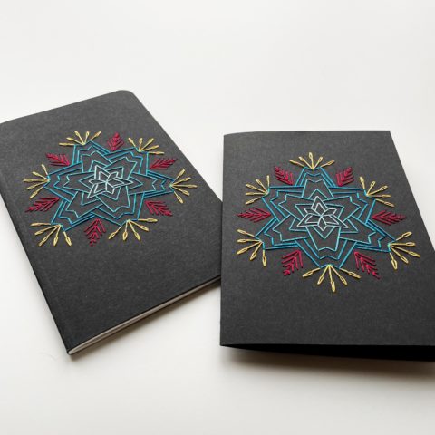 A star-shaped flower in blue, raspberry, and yellow, stitched on a black greeting card and notebook