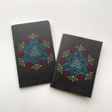 A star-shaped flower in blue, raspberry, and yellow, stitched on a black greeting card and notebook