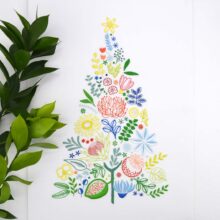 A multicolored embroidery on white of various leaves, fruits, and flowers arranged into a triangular tree shape with a star on top, shown next to an olive sprig