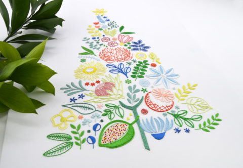 A multicolored embroidery on white of various leaves, fruits, and flowers arranged into a triangular tree shape with a star on top, shown next to an olive sprig and photographed at an angle