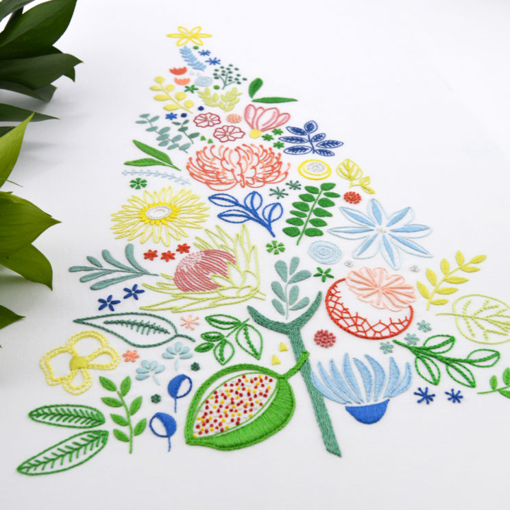 A multicolored embroidery on white of various leaves, fruits, and flowers arranged into a triangular tree shape with a star on top, shown next to an olive sprig and photographed at an angle