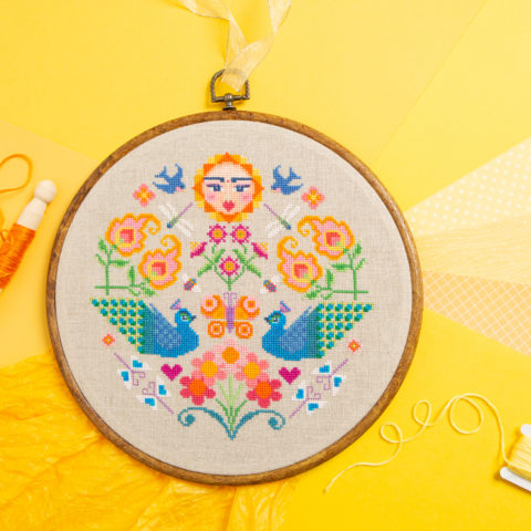 A colorful folk art cross-stitch showing birds, insects, and flowers with a sun shining down. Displayed in a wooden hoop on a bright yellow background