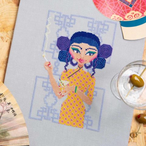 Cross-stitch of a woman in a yellow dress standing in front of a chinese screen holding a martini and a cigarette