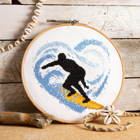 cross stitch of a black silhouette surfing on a gold board inside a blue, heart-shaped wave