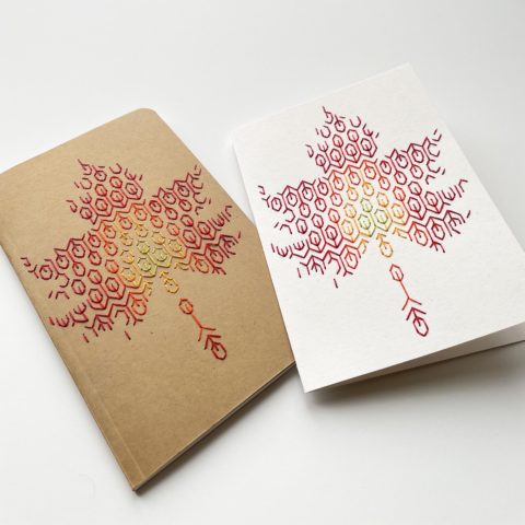 A blackwork leaf design in fall colors embroidered onto a kraft brown notebook cover and a white greeting card, viewed from a low angle on a white surface