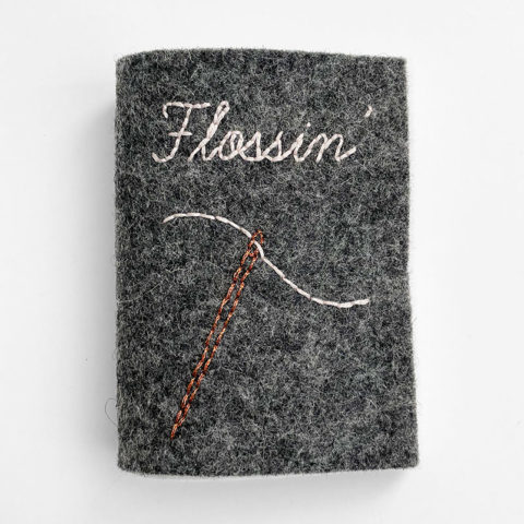 A small felt book in dark grey with an image of a needle and the word "flossin'" embroidered on the front