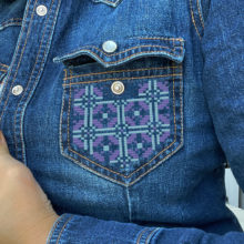 Geometric tatreez pattern stitched onto the pocket of a denim shirt in purple and light blue