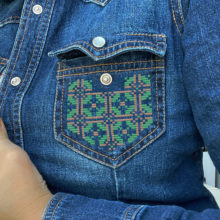 Geometric tatreez pattern stitched onto the pocket of a denim shirt in green and gold