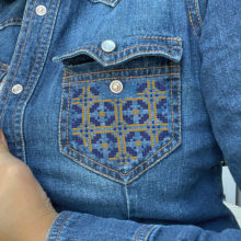 Geometric tatreez pattern stitched onto the pocket of a denim shirt in dark purple and gold
