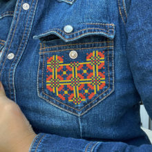 Geometric tatreez pattern stitched onto the pocket of a denim shirt in red and yellow