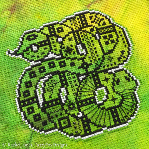 A cross stitch of a snake coiled around itself several times and filled with black folks art patterns on green hand dyed fabric