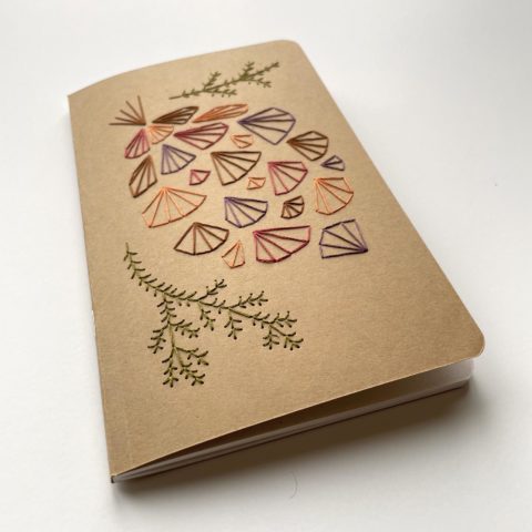 A geometric pinecone design in fall colors embroidered onto a kraft brown notebook cover, viewed from a low angle on a white surface
