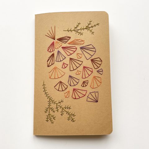 A geometric pinecone design in fall colors embroidered onto a kraft brown notebook cover