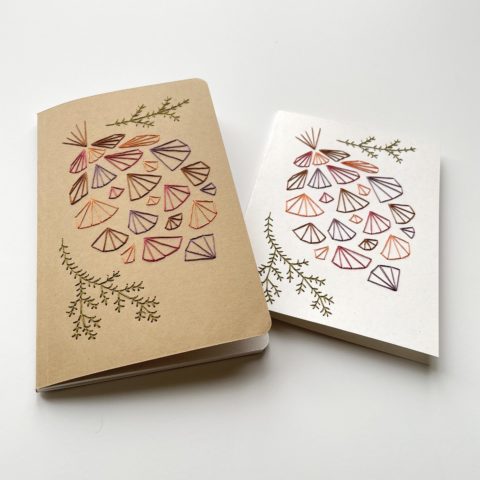 A geometric pinecone design in fall colors embroidered onto a kraft brown notebook cover and a white greeting card, viewed from a low angle on a white surface