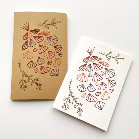 A geometric pinecone design in fall colors embroidered onto a kraft brown notebook cover and a white greeting card
