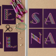 Purple postcards, each with one letter printed on it spelling out the words "heal" and "sanar," embellished with colorful geometric embroidery patterns and laid on a cork background
