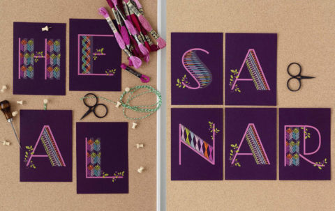 Purple postcards, each with one letter printed on it spelling out the words "heal" and "sanar," embellished with colorful geometric embroidery patterns and laid on a cork background
