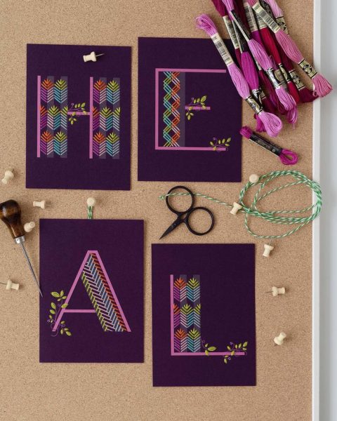 Purple postcards, each with one letter printed on it spelling out the word "heal" embellished with colorful geometric embroidery patterns and laid on a cork background