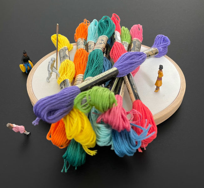 Tiny people standing, sitting, and climbing around a giant pile of embroidery supplies