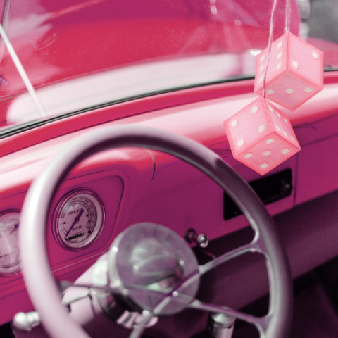 The dashboard of a pink classic car with two pink dice made of cross-stitch canvas hanging from the rear-view mirror