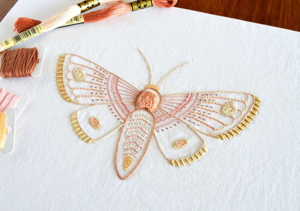 Anatomical embroidery of a moth in pinks and yellows on white fabric, with skeins and bobbins of floss to the left