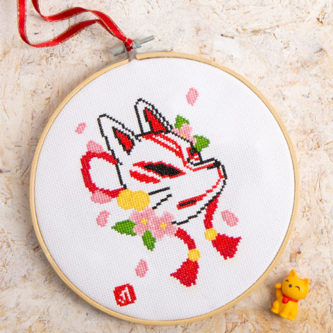 Cross-stitch design of a kitsune fox in a wooden hoop with a red ribbon