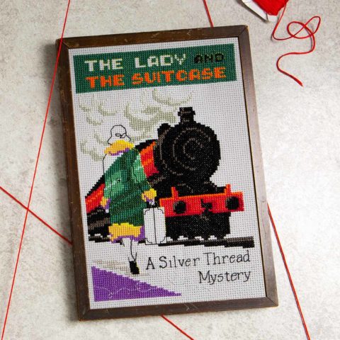 A cross-stitch of the mystery book cover The Lady And The Suitcase showing a brightly colored locomotive
