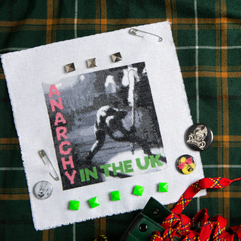 Cross-stitch of a greyscale image showing a musician smashing his guitar on stage with an overlay of the words "anarchy in the UK" in pink and green. It's displayed on a green plaid background with various British punk pins