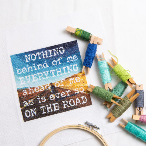 A cross stitch of the Jack Kerouac quote "Nothing behind me, everything ahead of me, as is ever so on the road" stitched over a picture of the open road with floss and hoop nearby