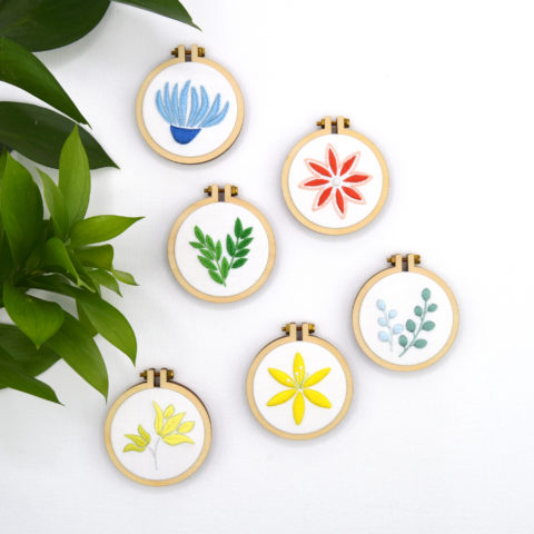 Six mini wooden embroidery hoops, each featuring a tiny embroidered botanical element in a single color on a white background, photographed with an olive sprig nearby
