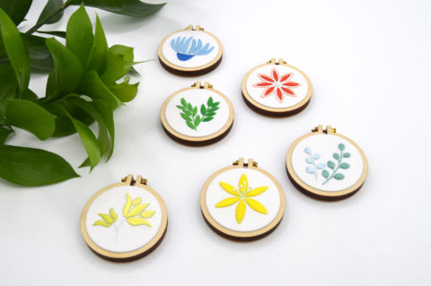 Six mini wooden embroidery hoops, each featuring a tiny embroidered botanical element in a single color on a white background, photographed from a low diagonal angle with an olive sprig nearby
