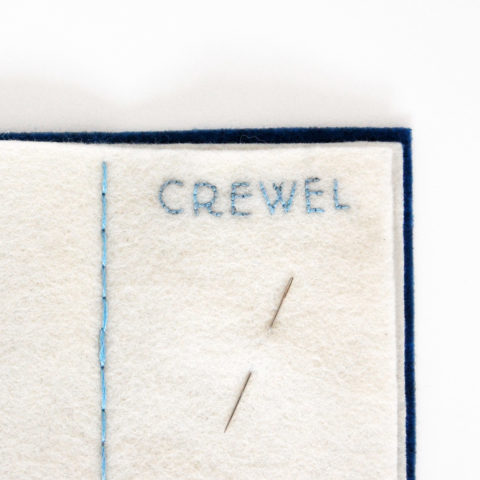 Close-up on the top of a Needle book page with the word crewel embroidered on it and a needle stuck in the fabric