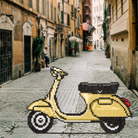 Cross-stitch of a yellow Vespa scooter superimposed on a photograph of a narrow Italian street