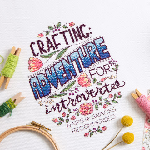 Cross-stitch of the words "Crafting: adventure for introverts, naps & snacks recommended" in various fonts with tulip ornamentation and stitching supplies scattered nearby