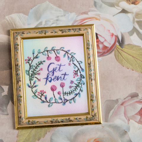 Cross-stitch of the words "Get bent" in purple cursive surrounded by a wreath of green leaves and pink flowers, displayed in a gold frame