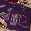Two purple postcards with the letters R and H printed on each embellished with colorful geometric embroidery patterns and laid on a cork background surrounded by stitching supplies