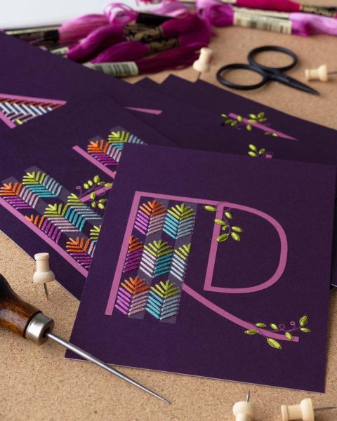 Two purple postcards with the letters R and H printed on each embellished with colorful geometric embroidery patterns and laid on a cork background surrounded by stitching supplies