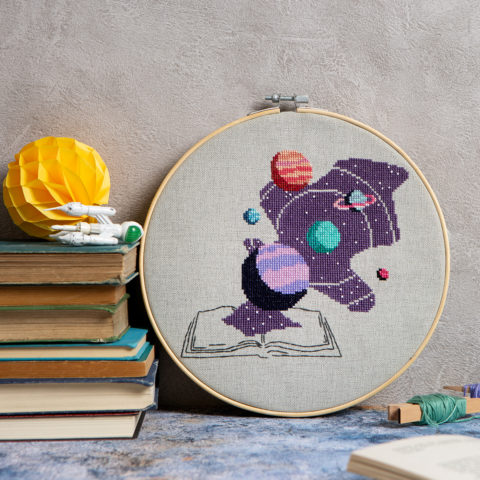A cross stitch next to a pile of books showing space and planets blooking out of the pages of an open book