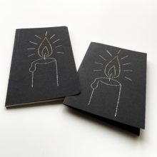 A drawing of a candle flame stitched on a black greeting card and notebook