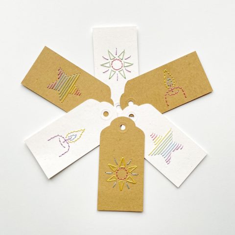 Three brown kraft paper and three white paper gift tags colorfully embroidered with an image of a star, a candle, and a sun