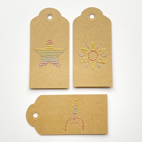 Three brown kraft paper gift tags colorfully embroidered with an image of a star, a candle, and a sun