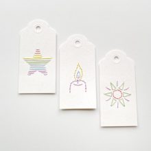 Three white paper gift tags colorfully embroidered with an image of a star, a candle, and a sun