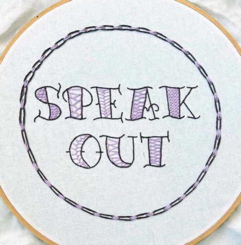 The words "speak out" embroidered in purple on white fabric in a wooden hoop, surrounded by a circular chain design