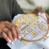 A close up of brown hands wearing two gold rings stitching a yellow Palestinian tatreez design in a wooden embroidery hoop