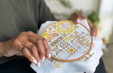 A close up of brown hands wearing two gold rings stitching a yellow Palestinian tatreez design in a wooden embroidery hoop