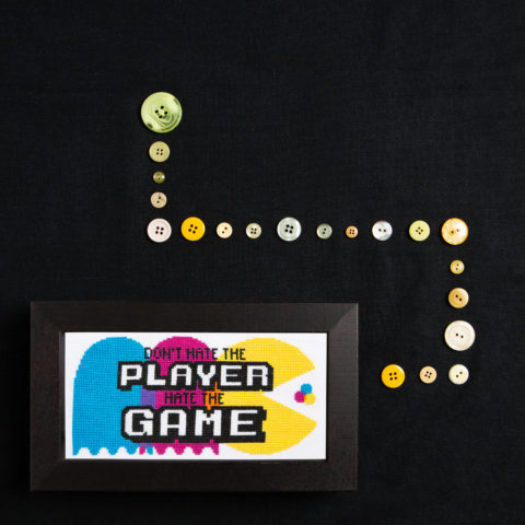 A cross-stitch of Pac-Man and two ghosts in CMYK colors overlaid with the words, "Don't hate the player, hate the game," displayed next to lines of buttons set out at right angles on a black background as though Pac-Man is about to eat them